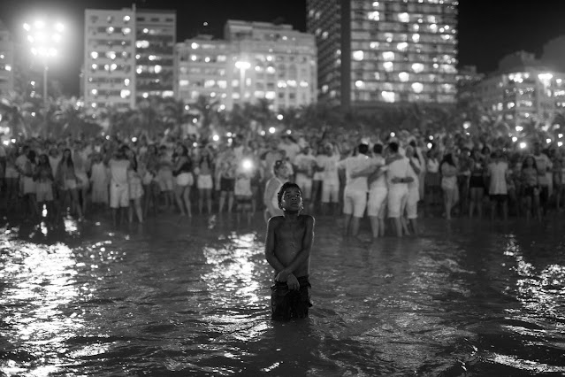 Garoto negro, sem camisa e com os braços cruzados, observa algo no céu. Ele está no mar, com as águas batendo no joelho e ao fundo da imagem, distante do garoto e na areia da praia, pessoas vestidas de branco se abraçam e tiram fotos.