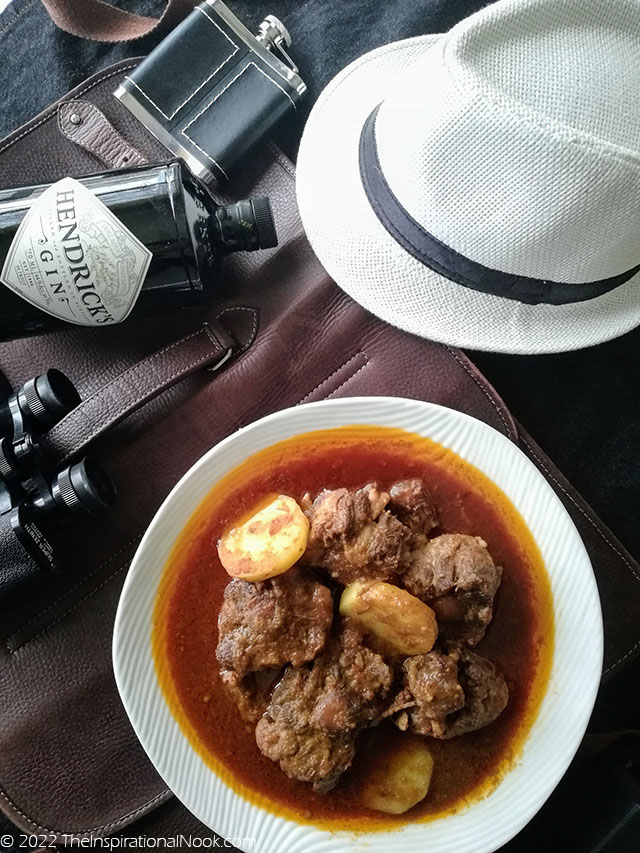 A plate of beef oxtail curry and potatoes on a leather bag with binocular, a bottle of gin, a hip flask and a white hat