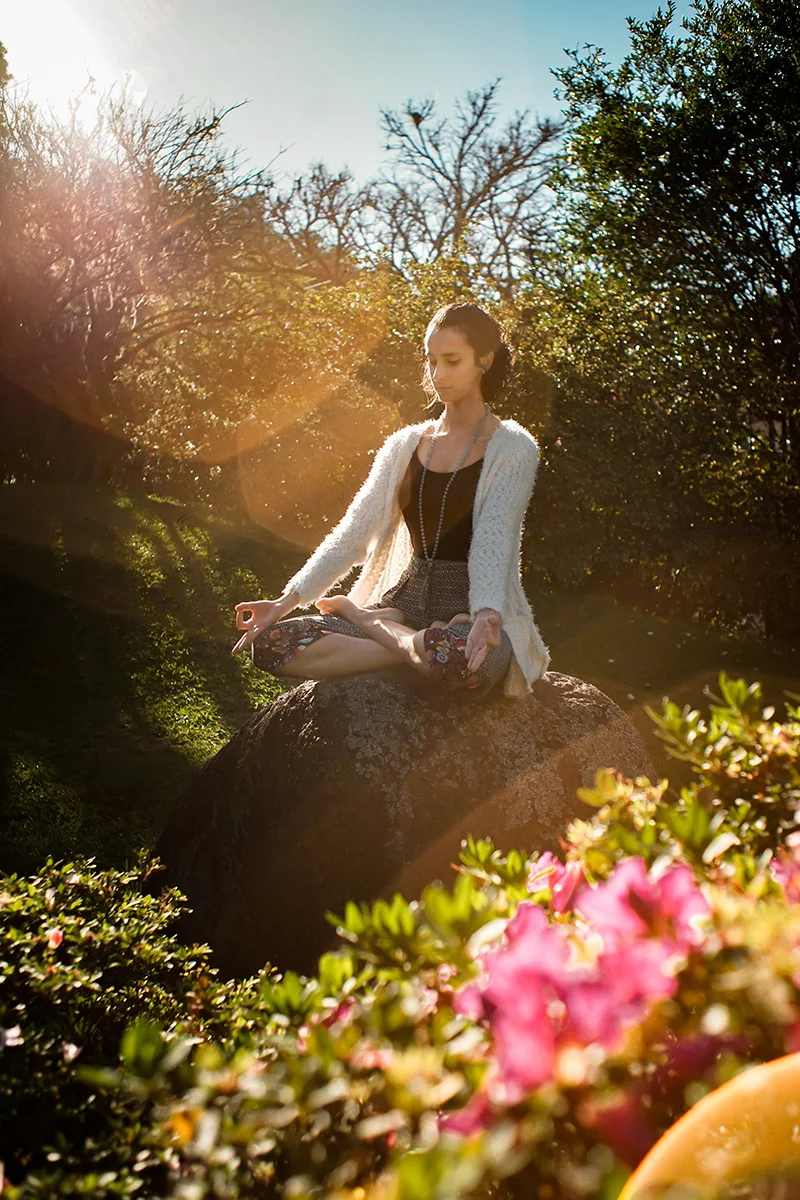 young woman in cardigan and a messy bun hair is meditating in a flowering garden