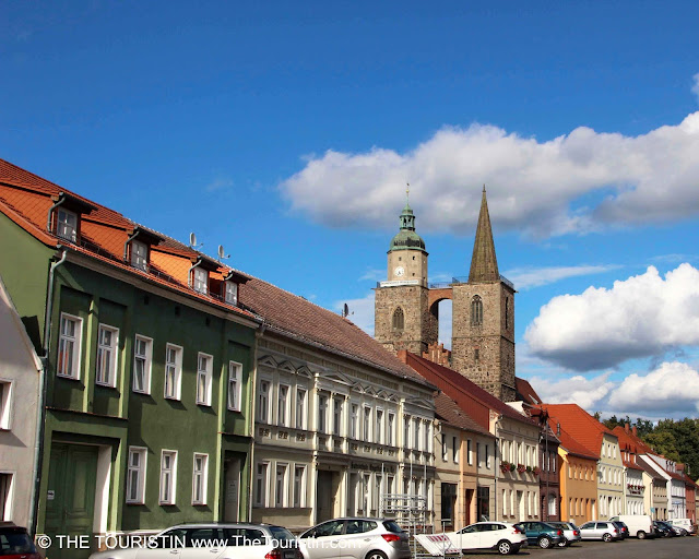 Cars park in front o a row of colourful houses in front of a church with two spires under a blue sky.