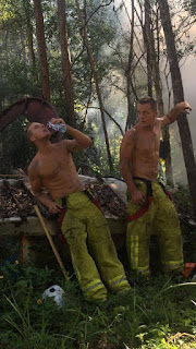 Foto(uma das fotos do calendário 2017 Austrália). Dois bombeiros desnudos da cintura para cima em meio a selva com muita fumaça branca; usam calças verdes de tecido grosso com bolsos chapados na altura das coxas, listras duplas em verde-limão nas laterais e na altura dos tornozelos, cinto com tiras largas de segurança em preto e vermelho. Eles têm porte atlético, sarados, músculos fortes e abdomens definidos. Estão em pé, lado a lado, um, à esquerda, apoia o bumbum e o antebraço direito em um pequeno barco abandonado cheio de folhas secas dentro, com a mão esquerda segura uma garrafa de água próximo a boca, com a cabeça inclinada para trás, bebe sem encostar o gargalo; o outro, apoia o antebraço esquerdo em um fino e alto tronco enquanto observa o colega hidratando-se. Próximo a eles, há instrumentos e acessórios de combate ao fogo.