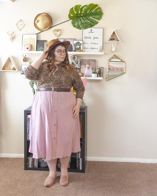 An outfit consisting of a brown panama hat, an animal print blouse tucked into a muted pink button down midi skirt and muted pink pointed mules.