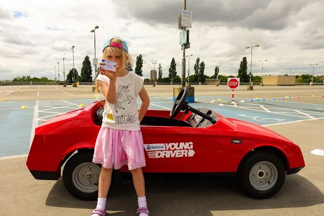 My daughter standing with attitude and holding her drivers license in front of a red two seater open top car with "Young Driver" on the side