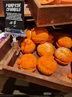 A photo of a large light brown square box containing a bunch of pumpkin shaped light orange bubble bars with white cream in the middle  next to a black rectangular card that says pumpkin crumble bubble bar in white font on a light brown rectangular table on a bright background