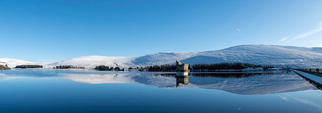 Backwater reservoir, water tower right of centre a jet trail in the sky reflected in the still water, blue skies and snow covered hills surround