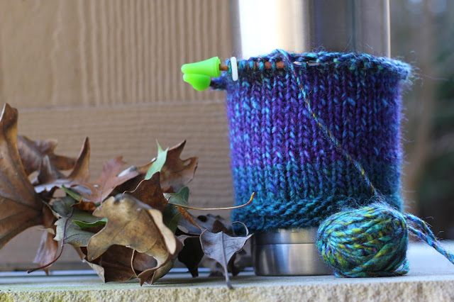 Jewel-toned knitting work in progress on circular knitting needles with green stoppers at the end of the needles to keep the work from sliding off. It's on a stainless steel thermos to try on for size. A small ball of yarn is attached to the project. It's sitting next to dry brown oak leaves.