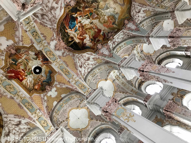 An elaborately decorated ceiling painting with a groups of people dressed differently in coloured robes and tunics, painted in pastel colours ranging from pink, light blue, and yellow, to red to gold.