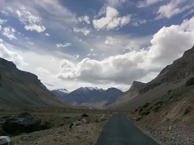Snow-capped Himalayan mountains partially covered in clouds, with a rocky valley and a narrow trail running through the remote Spiti region in Himachal Pradesh.