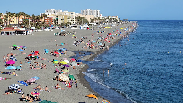 Vista aérea de una gran playa con mucha gente, edificios a su espalda y las azules aguas del mar a su frente.