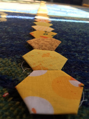 A chain of seamed-together yellow fabric hexagons, all different prints and shades, that stretches into blurry distance, laid flat on a sunlit blue and green carpet.