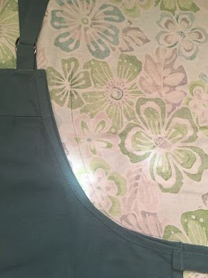 A close up of the bib of a colonial-blue cooking apron, with neck strap arranged neatly pointing upward and long straps vanishing out of frame to the right. A faint pen line arcs up the faintly patterned back of the green, turquoise, and grey tropical floral fabric from the right edge of the apron bib, separating from the edge of the bib as it curves upward, ending even with the top edge but about an inch further to the right.