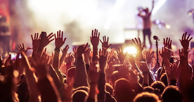 A large crowd at an outdoor festival with their hands raised in the air. In the distance, there is a person on stage.