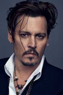 Foto em close do ator Johnny Depp, um moreno claro com rosto em formato quadrado, cabelos curtos lisos com franjão, quatro mechas bem fininhas desalinhadas com gel descem sobre o olho direito até quase a altura dos lábios, as sobrancelhas retas emolduram os olhos focados diretamente para nós, em formato amendoado castanhos escuros com uma linha fina de lápis preto nos cílios inferiores, o nariz é fino e reto, o bigode tem fios ralos escuros, voltados um pouco abaixo dos lábios médios, rosados e bem desenhados, o cavanhaque acompanha a linha do bigode mas não se une à ele, tem raros fios brancos e detalhe de alguns pelos em formato de T abaixo do lábio inferior. Brincos de pequenas argolas prateadas adornam as orelhas do moreno, duas na orelha esquerda e uma na direita. A gola da camisa branca despojadamente voltada para dentro deixa à mostra abaixo dela parte uma corrente de elos prateados sobre blazer ou algo do gênero, em preto. No peito do ator, um colar curto estilo tribal, com pequenos canudinhos de madeira intercalados com contas claras e em tons de marrom finalizado com pingentes e detalhes em prata.