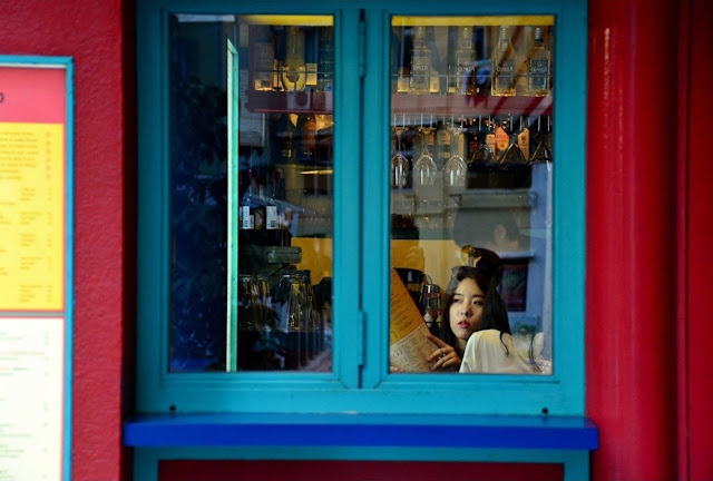 Através de uma janela, se vê uma mulher em um ambiente que parece um bar ou restaurante.