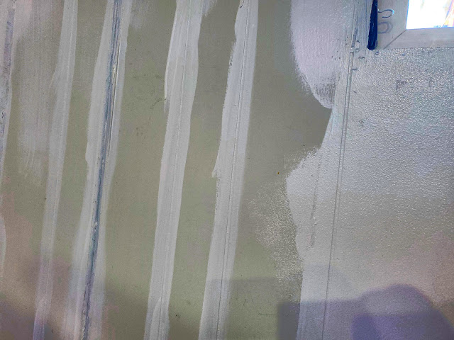 Vertical and diagonal streaks of white and grey paint overlap on a wall, suggesting an unfinished painting or renovation process. Shadows cast onto the surface hint at objects or people just out of view.