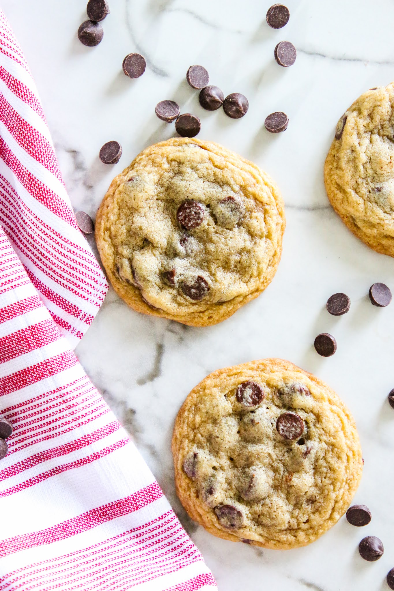 Three chocolate chip cookies on a marble table with scattered chocolate chips and a red towel