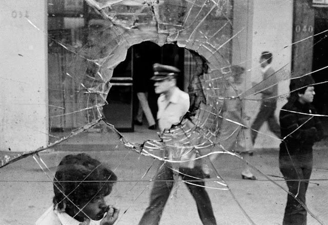 A fotografia em preto e branco mostra um vidro com um buraco no meio e, por isso, está trincado. Por meio do buraco pode-se ver o busto de um policial andando na rua. Há outras pessoas na rua, porém o foco é o policial e o buraco no vidro.