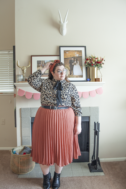 An outfit consisting of a rose colored rose headband, a brown and black animal print collared shirt with a black tie tied into a bow, tucked into a rose pleated midi skirt and black chelsea boots.