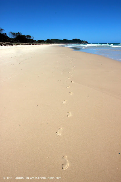 Footsteps on the beach with green hills and the ocean in the background under a cloudless blue sky..