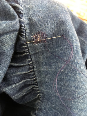 A needle with blue-purple thread woven through a loose lattice of blue-purple thread covering a small hole in a piece of denim fabric, just above a gathered seam.