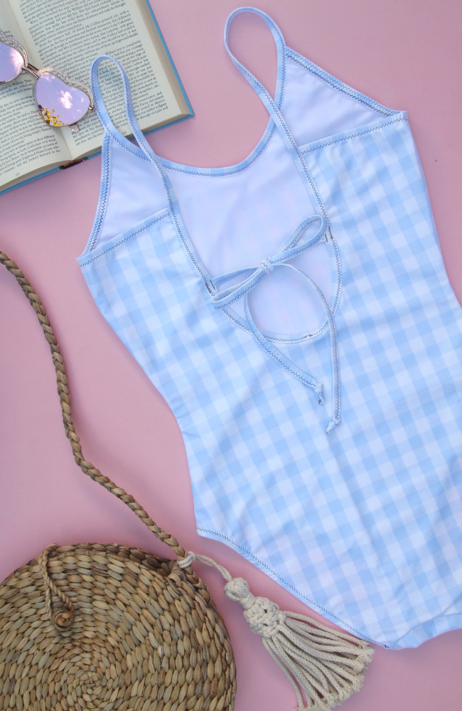 A light blue and white gingham swimsuit with a bow tie back on a pink background, with a straw bag and sunglasses