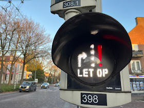 Lamp bij verkeerslicht met rood uitroepteken, tekst “let op” en schetsje dat waarschuwt voor overstekers