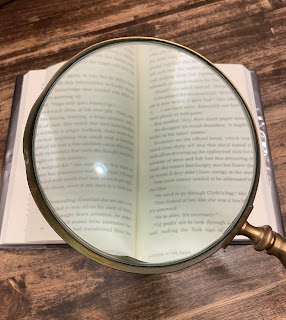 an open book on a wood grain table with a large magnifying glass held over it so the center of the words are large.