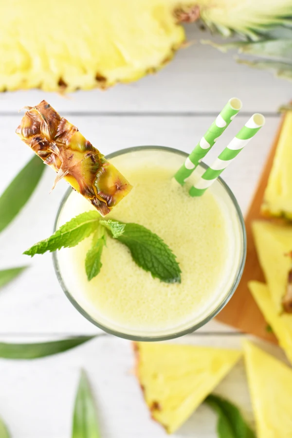 Top view of a tall glass filled with a refreshing pineapple banana smoothie, featuring a pineapple triangle garnish, two striped green straws, and fresh mint leaves.