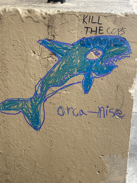 Killer whale drawn partially over graffiti that says "Kill all commies," word "commies" covered and replaced with "cops," "Orca-nise" written under whale