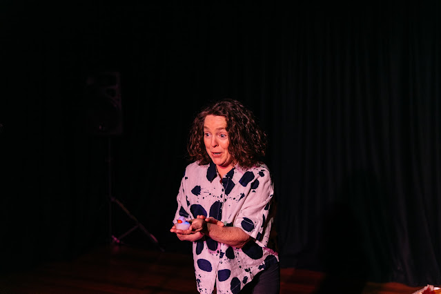 Poet and performer Caroline Reid is on stage at The Goodwood Theatre Studio, performing a scene from her spoken word theatre show SIARAD. She wears a bold cowprint button shirt and holds a tiny rubber duck in her hands. Her face expresses surprise and delight.