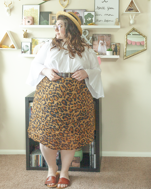 An outfit consisting of a straw boater hat, a white off the shoulder blouse with bell sleeves tucked into a brown leopard midi skirt and brown slide sandals.