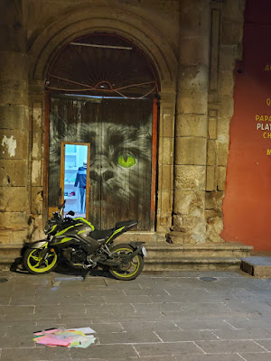 a night shot of a motorcycle parked in front of a shoe shop with an open door with a large face of a black cat painted around it. there are a pile of papers in the foreground.