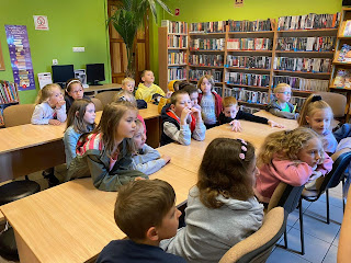 Tło: sala biblioteczna. Grupa dzieci przedszkolnych siedzi przy stolikach.