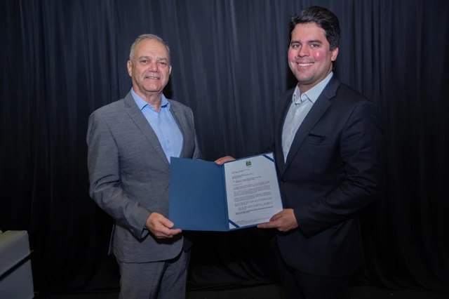 Na esquerda, com terno cinza, Paulo Wanderley (homem branco com cabelo branco) segura uma placa junto de André Fufuca (homem branco, moreno e com as bochechas rosadas) segurando a parte com papel da placa e está sorrindo. Ele está de terno preto