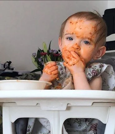 A baby eating spaghetti in a tomato based sauce with her hands