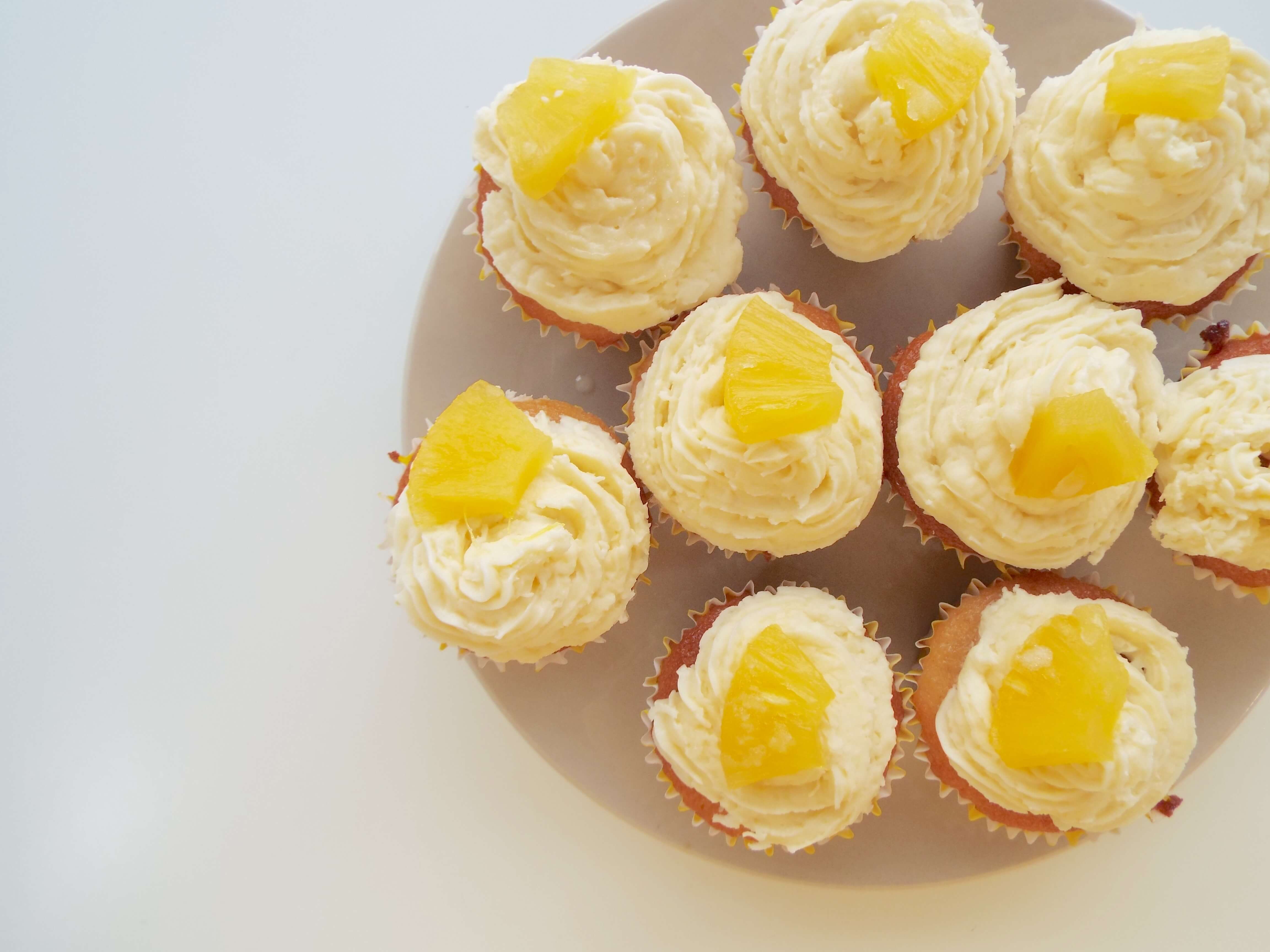Plate of nine, coconut cream buttercream iced cupcakes, with a pineapple slice on top of each.