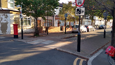 A side road junction where the surface has been raised to footway level with a gap running through for cycles. There are traffic signs, trees and houses in shot.