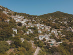 The photo shows a mountain village with white houses and trees. The village is located on a mountain slope, and other mountains can be seen in the distance. The sky is blue and cloudless.