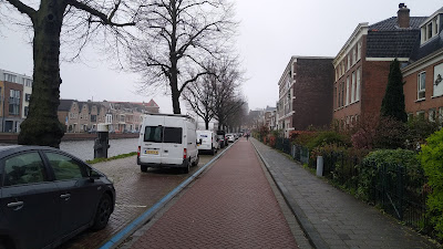 A narrow road with parking left and houses right beyond a footway. There is a canal to the right of the parking.