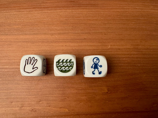 three dice with pictures on them on a wooden table. The pictures are a hand, waves, and a stick-figure person walking.