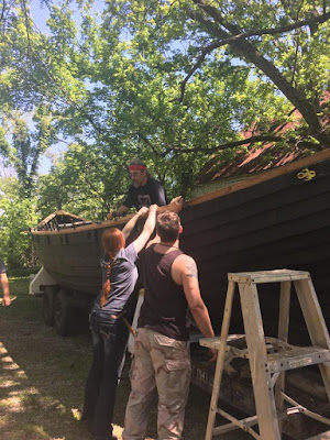 A clinker-built Viking ship on a trailer in sun-dappled  clearing, with a bright new rail clamped on, a man working on the rail from inside the boat, and a redheaded woman and another man standing outside th eboat, holding the rail in place.