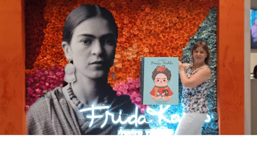 Kobieta z książką z serii Mali Wielcy pod tytułem Frida Kahlo na tle plakatu reklamującego wystawę Frida Kahlo. Życie ikony.