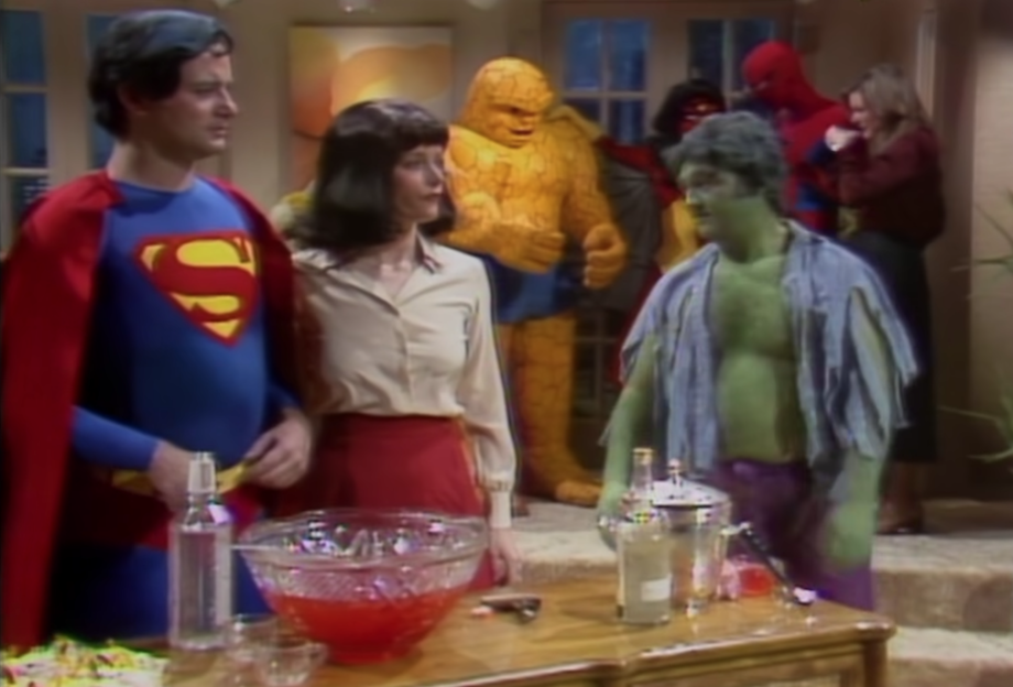 Bill Murray as Superman, Margot Kidder as Lois Lane, and John Belushi as Hulk at beverage table with other superheroes in background