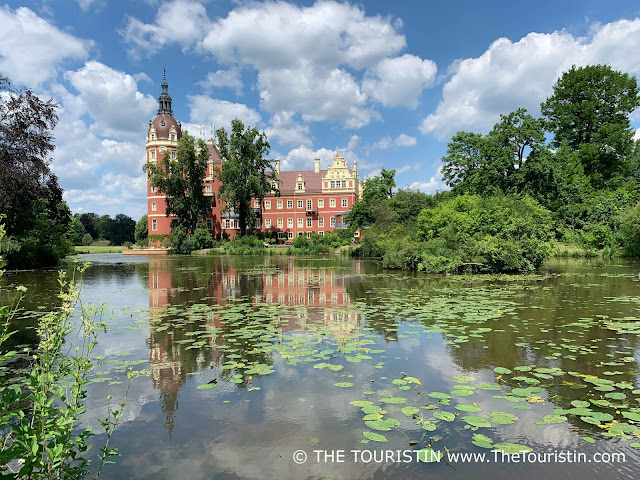 Water lilies on a lake in front of a red-ish coloured renaissance castle under a blue sky with fluffy clouds.