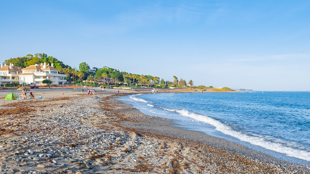 Playa de arena con piedras, el mar azul a su frente y edificios con vegetación al fondo.