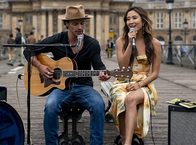 Un hombre toca la guitarra y una mujer canta con un micrófono a su lado. Ambos están sentados.