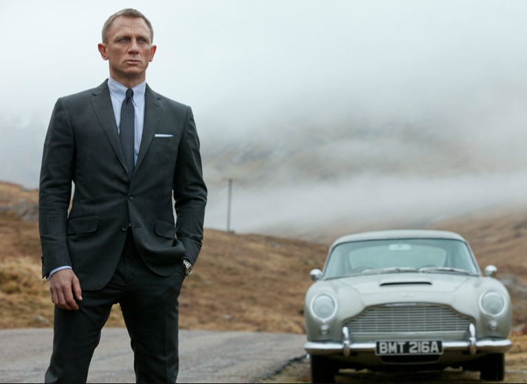 Daniel Craig as Bond standing by the side of the road next to the Aston Martin
