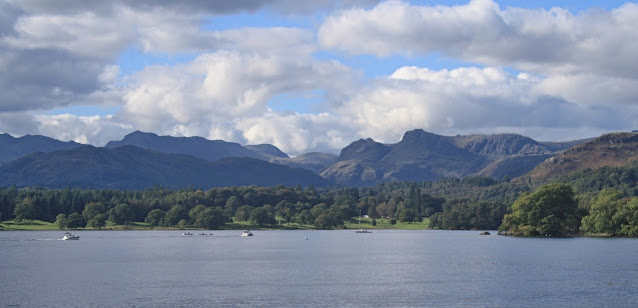 A view of boats on Lake Windermere, with hills in the background. It's a sunny day with some, scattered clouds.