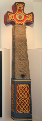 A tall thin stone cross shaft with carved looping patterns. The cross and base have been recreated and are painted in blue, red and yellow.