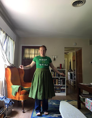 A young redheaded woman in a living room crowded with furniture and sewing supplies, facing the camera and smiling with arms outstretched. She's wearing a pleated green skirt over dark jeans and a kelly green t-shirt that reads "keep it real."